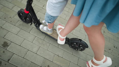 a close-up leg view of a woman and child standing on a scooter as the woman uses her leg to move the scooter forward on an interlocked path