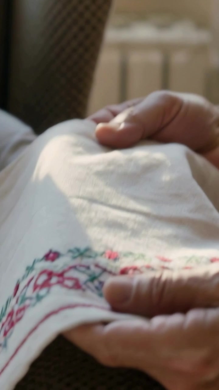 Vertical video: Pulling back to man in brown sweater smoothing embroidered hem at window