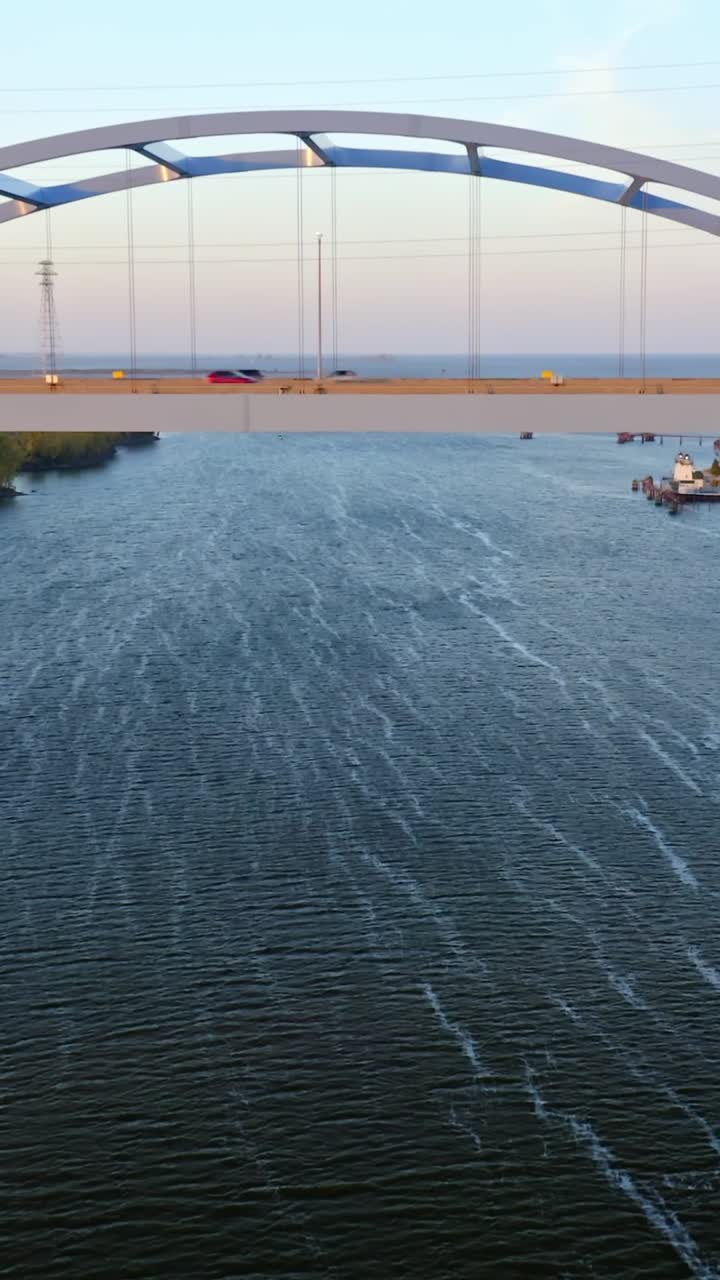 Vehicles move steadily across a sleek arch bridge spanning a wide river, while gentle waves shimmer below in warm evening light creating a tranquil, balanced scene