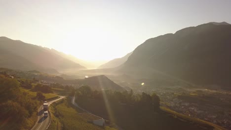 Drone-shot-in-the-early-morning-with-the-sun-rising-over-a-beautiful-mountainous-valley-with-some-traffic-on-the-road-in-Lavaux-Oron,-Switzerland-in-4k