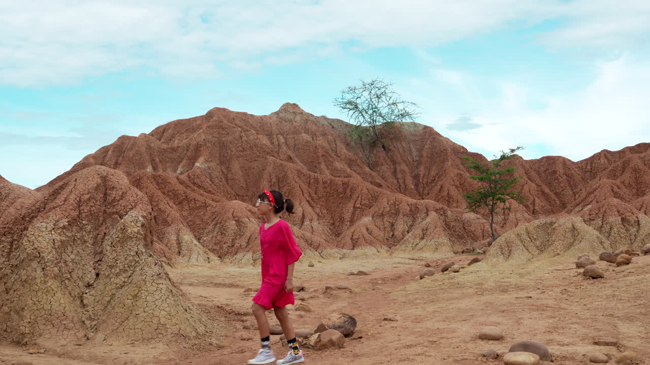 A young child in a vivid red outfit explores the rugged, arid terrain of Tatacoa Desert under a clear blue sky, with dramatic rock formations and scattered vegetation enhancing the scene.