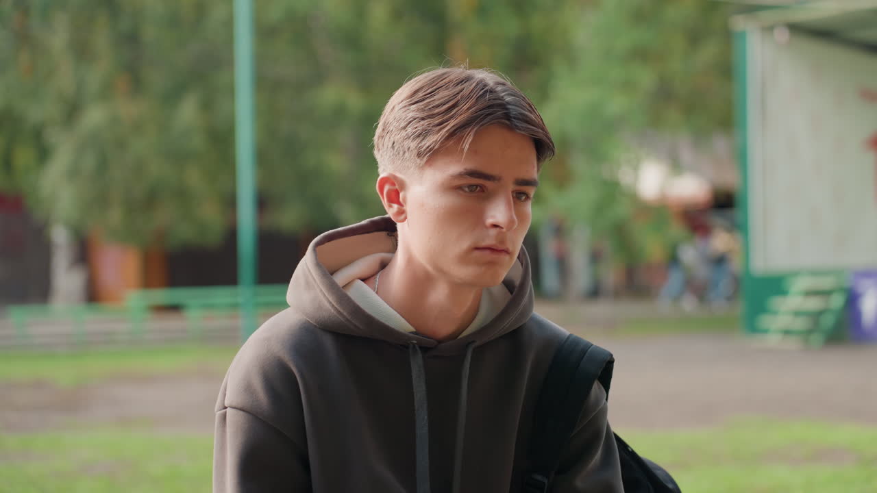 adolescent pensif en sweat à capuche, portrait sur un banc de parc capturant une ambiance introspective, fond bokeh doux et lumière chaude du soleil sur le visage, jeune étudiant attendant avec un sac à dos, cadrage cinématographique mettant l’accent sur la subtilité