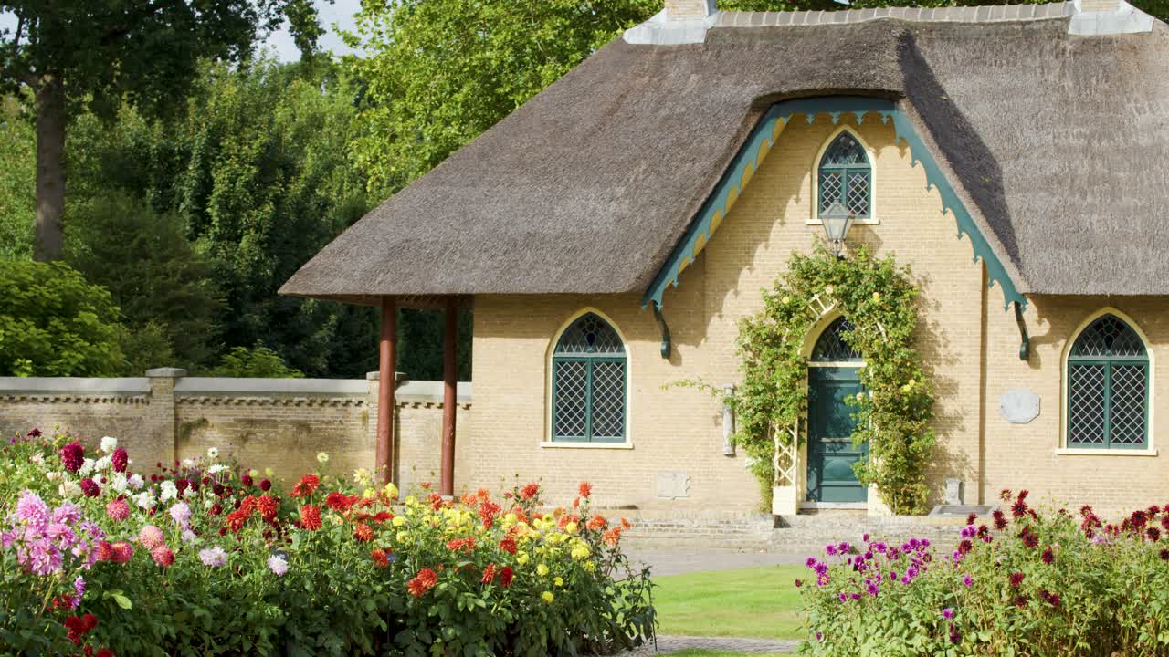 Camera slowly pans past thatched cottage and vibrant dahlia flower beds in bright daylight