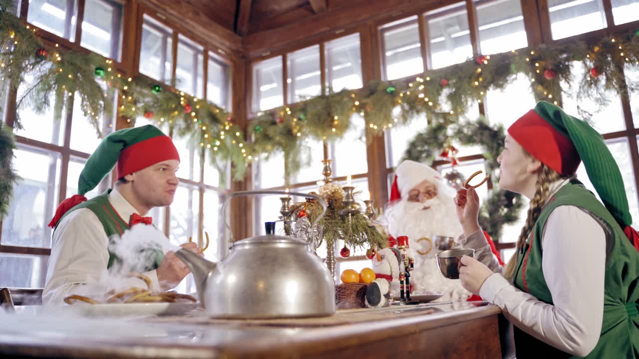 Happy elves and Santa at the table on Christmas decorations background. Fairy characters and Santa drinking tea with cookies on New Year's time.