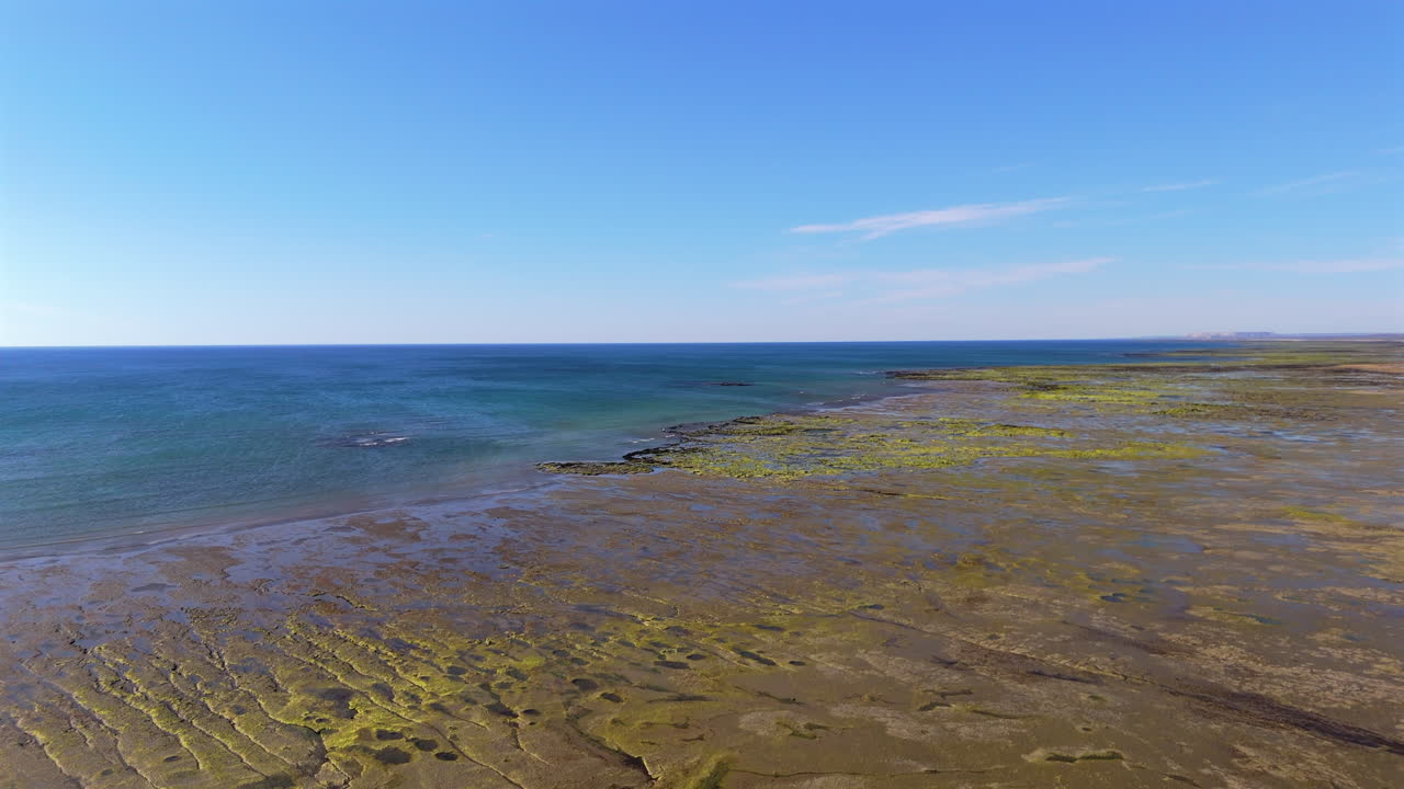 An aerial view of Las Grutas at low tide, showcasing the sandy shoreline and exposed rocky formations. The blue ocean contrasts beautifully with the dry land