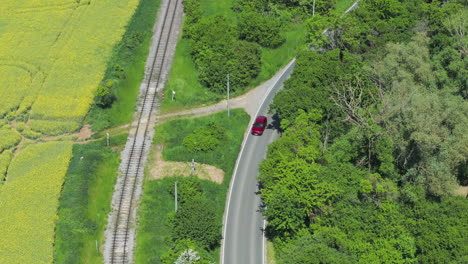 drone shot of red car on countryside road by railway and green landscape, trees and meadows