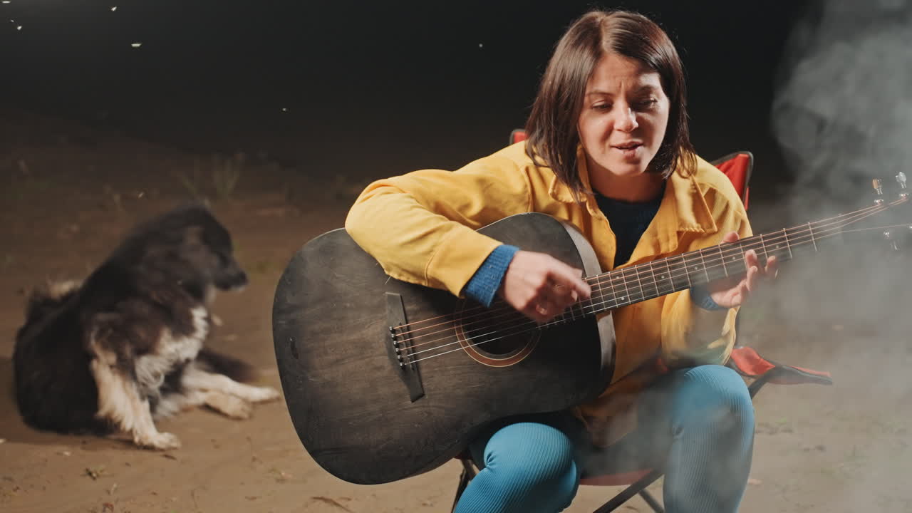 Donna con chitarra e cane che cantano. Donna e cane rilassati si godono la musica intorno al falò di notte. Ambiente esterno sereno con donna che strimpella la chitarra e cane accanto al fuoco sotto il cielo stellato.