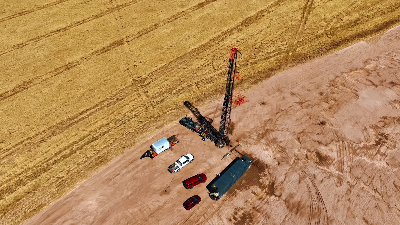 Flying above around the rig, few trucks and cars in the vast field. Wheat field with ripe crop nearby.