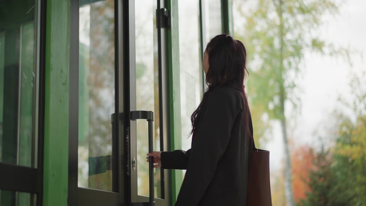 Woman in black blazer and white pants walks through outdoor area with green structural elements holding phone. Woman carries brown bag while moving past trees showing autumn foliage