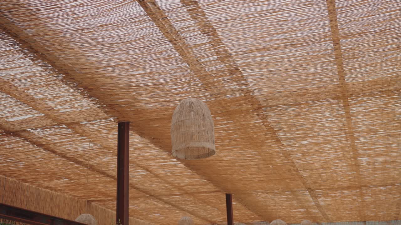 Vista verso l'alto di un baldacchino in paglia intrecciata a soffitto con lampada in vimini sospesa che crea una calda texture naturale e un morbido motivo di luce.