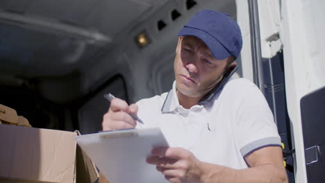 focused courier calling customer in van and writing on clipboard