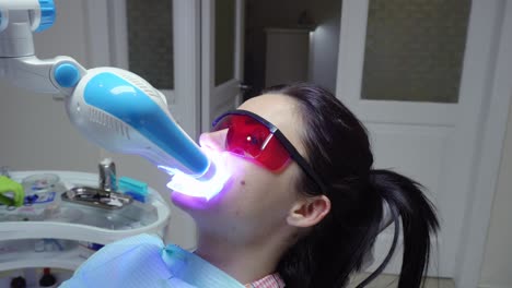 Young-woman-with-an-expander-in-mouth-and-red-protective-glasses-getting-UV-whitening-at-the-dentist's-office-by-an-ultra-violet-machine.-Shot-in-4k