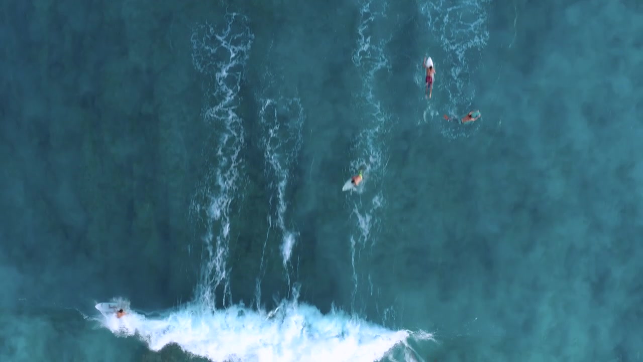 Droneoptagelse ovenfra af surfere i køen, mens en surfer rider på en stor brydende bølge, der sender hvidt skum hen over revet, og fanger både gruppen og bølgens dynamiske energi.