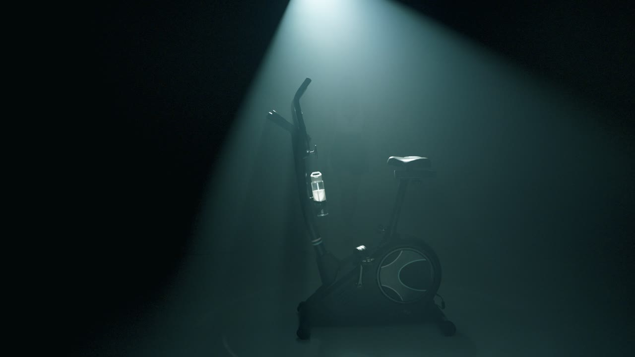 Profile shot of an empty stationary bike glowing in cinematic backlight within a smoky, dark studio. A woman steps from the shadows, mounts the bike, and starts her workout with focused energy