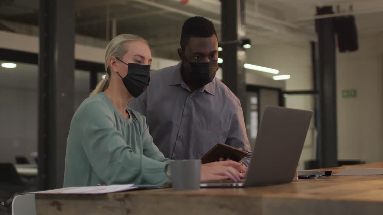 Diverse male and female business colleagues wearing masks in discussion at work using laptop