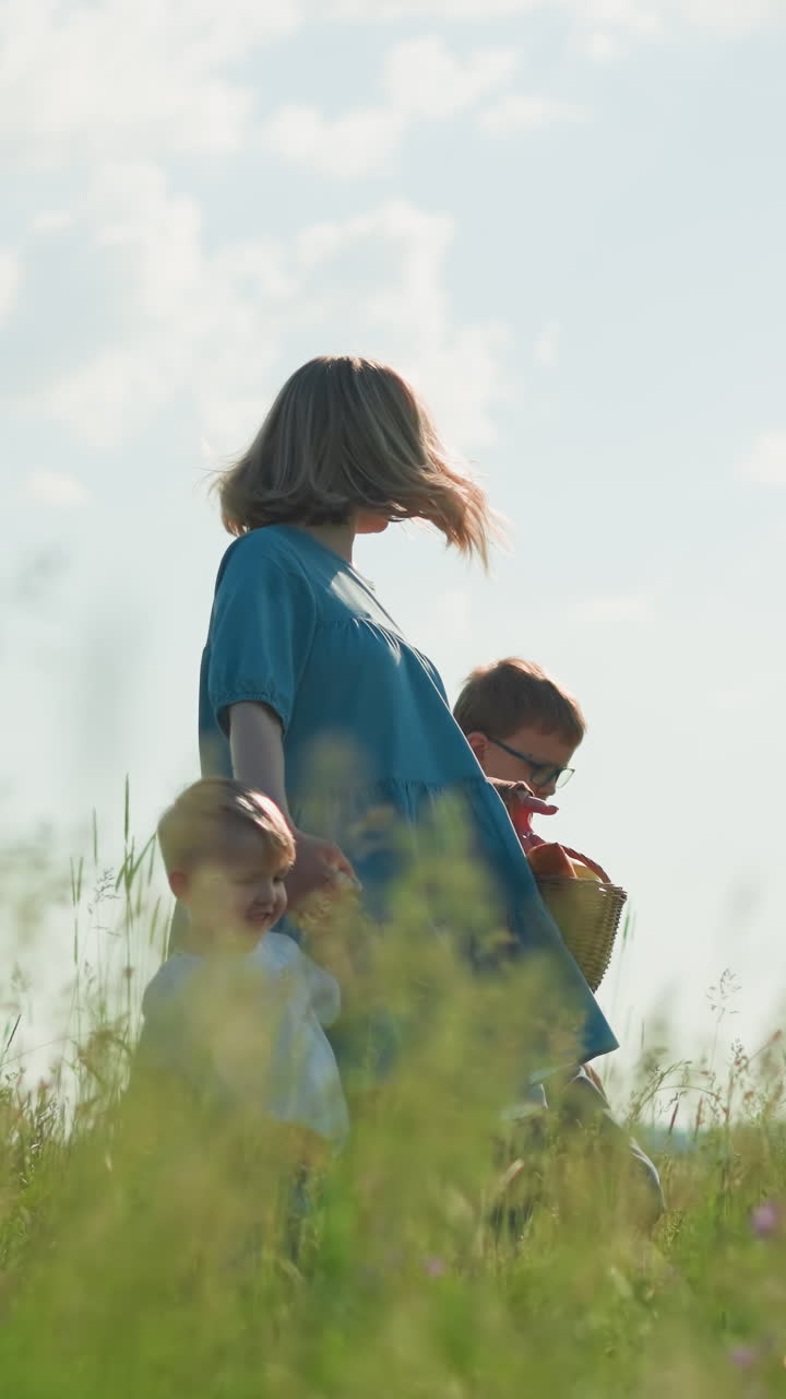 A joyful woman in a flowing blue gown walks through a lush green grass field with her two young sons, both in white shirts. She carries a basket, and they all smile under the clear blue sky