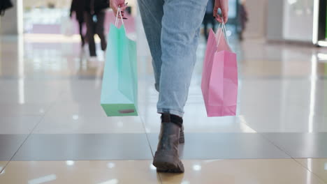 lower body view of person walking in sleek mall tiles while carrying two shopping bags, wearing jeans and black boots, with other shoppers moving in the background in a modern mall environment