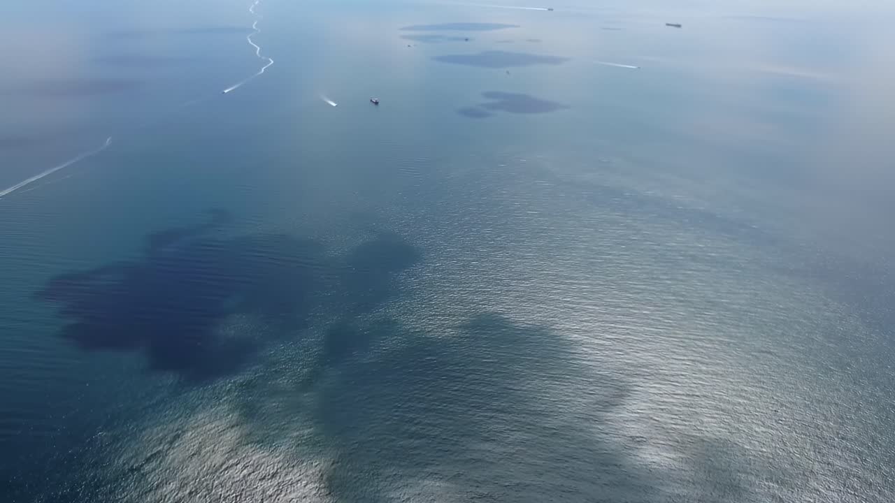 This captivating aerial view highlights various textures in the ocean as boats cruise across the calm waters under a clear sky. Sunlight reflects beautifully on the surface creating patterns.