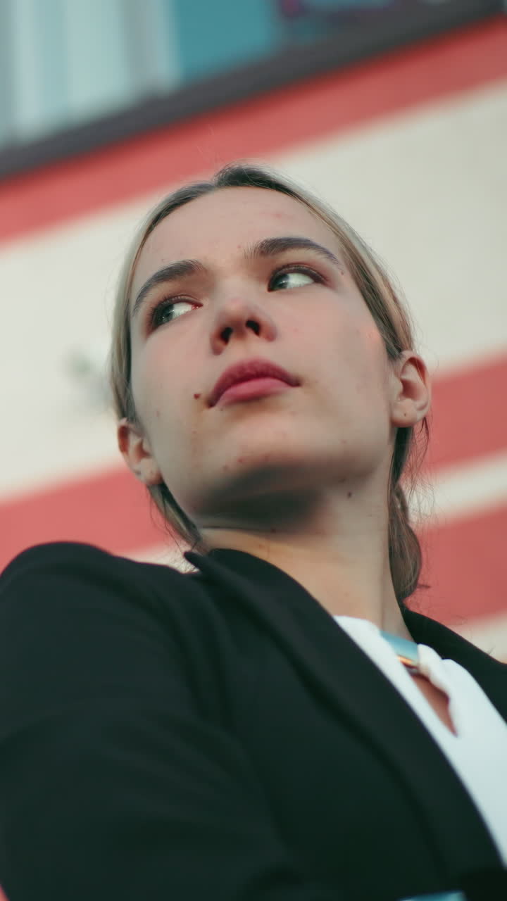 Young damsel in black blazer stands with arms crossed, gazing into distant sky with confident expression in front of modern curved building