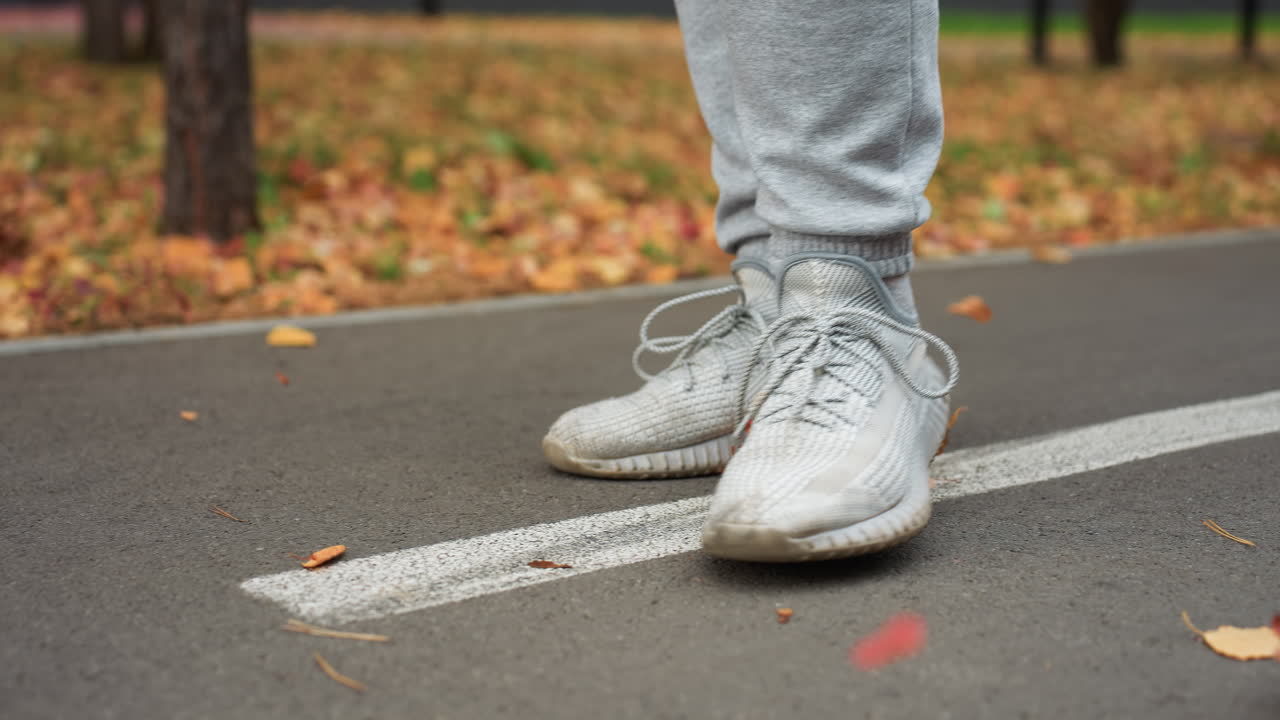 Lower angle view of person walking along tarred road as autumn leaves move with wind, some stepped on, surrounded by fallen foliage and tree-lined path in serene park environment on breezy fall day