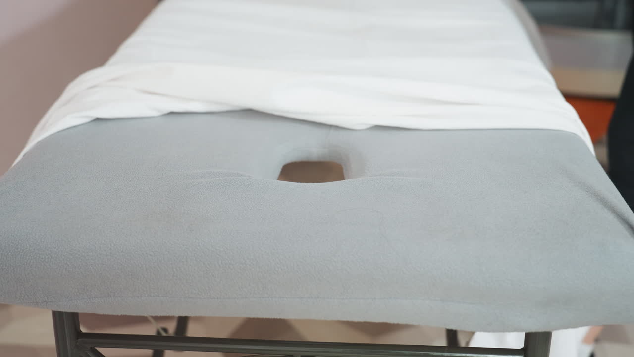 Woman spreading white sheet over soft gray massage bed supported by black metal stand, preparing surface in calm, clean setting with focus on neatness, fabric texture, and professional readiness