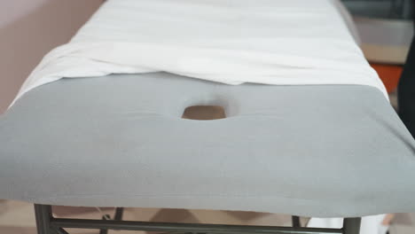 woman spreading white sheet over soft gray massage bed supported by black metal stand, preparing surface in calm, clean setting with focus on neatness, fabric texture, and professional readiness