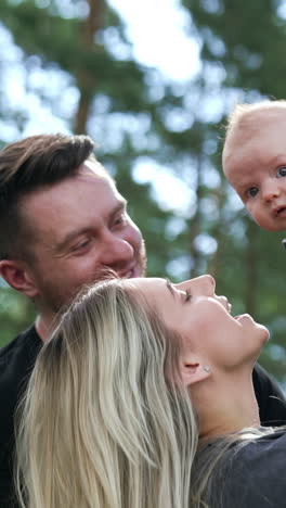 happy blonde woman holding her newborn baby over her head. bearded man stands close looking at child with love. nature backdrop. vertical video