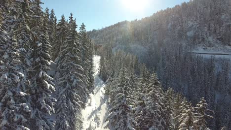 Flying-over-a-snowy-forest-path-with-a-ski-resort-in-background.-Sunny-day