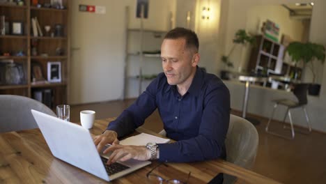 and adult man is sitting at a laptop. and adult man takes a sip of coffee and leans back in his chair