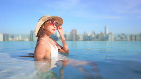 Elegante-Mujer-Asiática-En-La-Piscina-De-La-Azotea-Sobre-El-Horizonte-Urbano-Moderno-Disfrutando-Del-Sol-De-La-Tarde