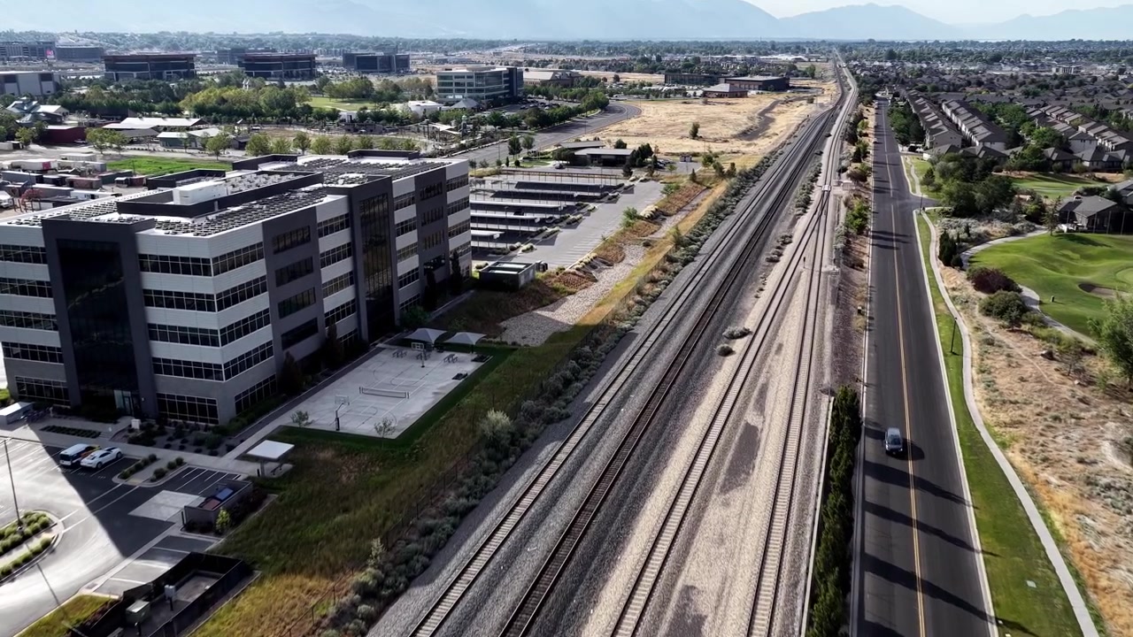 Vuelo de dron sobre edificios de oficinas modernos en Lehi, Utah, con tejados de paneles solares y estacionamiento solar cubierto junto a las vías del tren. Luz diurna brillante, paisaje urbano.