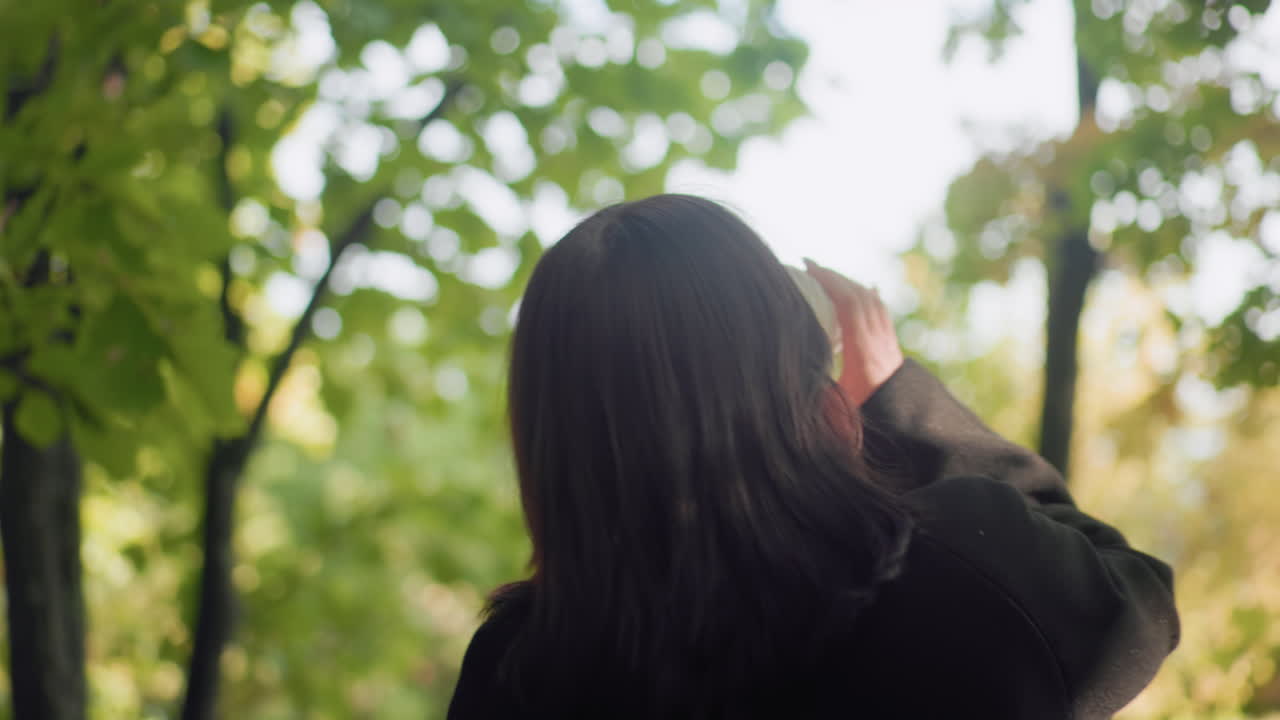 Medium back view of autumn lover strolling under leafy canopy, sipping warm drink with unhurried steps, black jacket and boots moving along forest path as sunlight filters through leaves