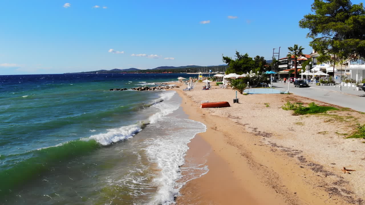 Aerial drone view of the Aegean sea coast in Nikiti, Greece. Beach with waves of the sea, umbrellas, sunbeds greenery along the embankment street, multiple buildings