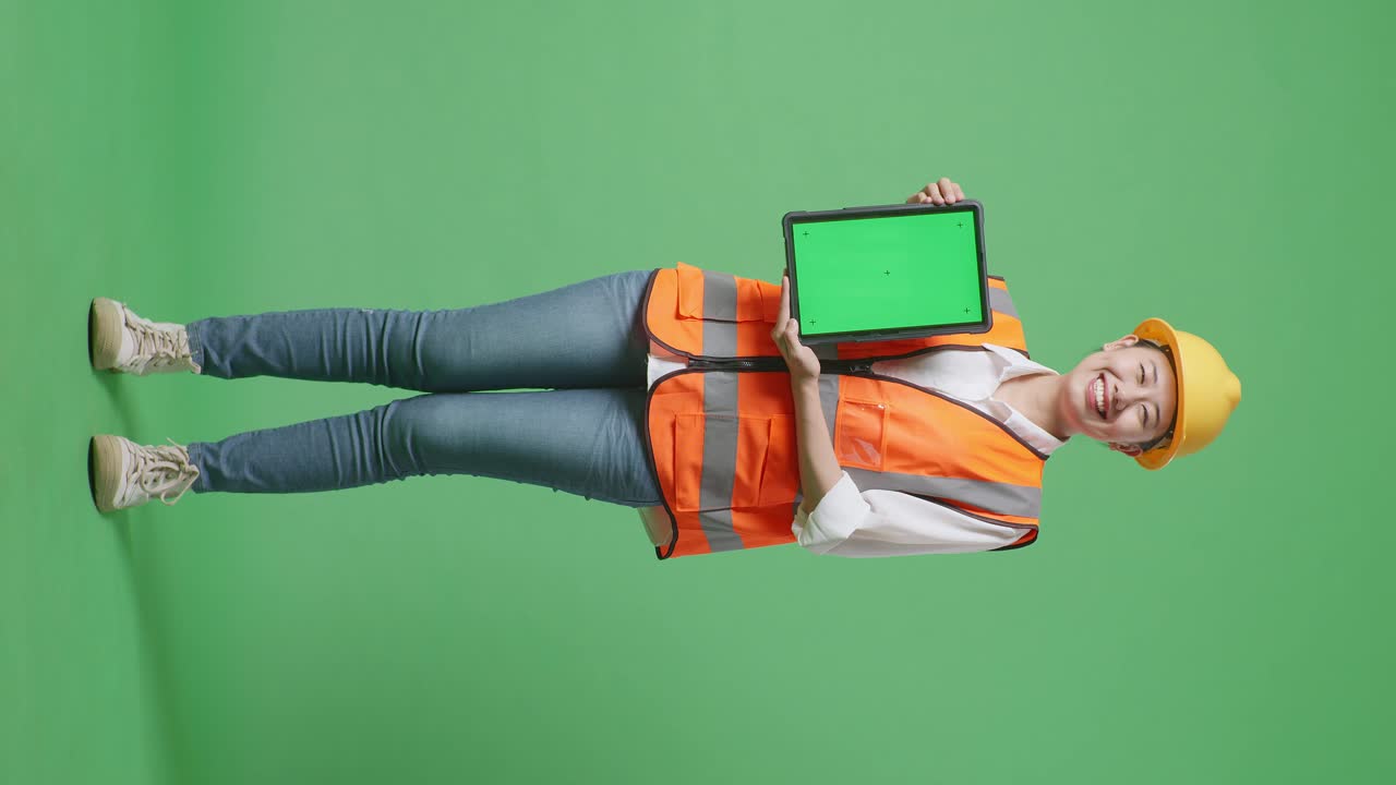 cuerpo lleno de ingeniera asiática con casco de seguridad sonriendo y mostrando la tableta de pantalla verde a la cámara mientras está de pie en el estudio de fondo de pantalla verde