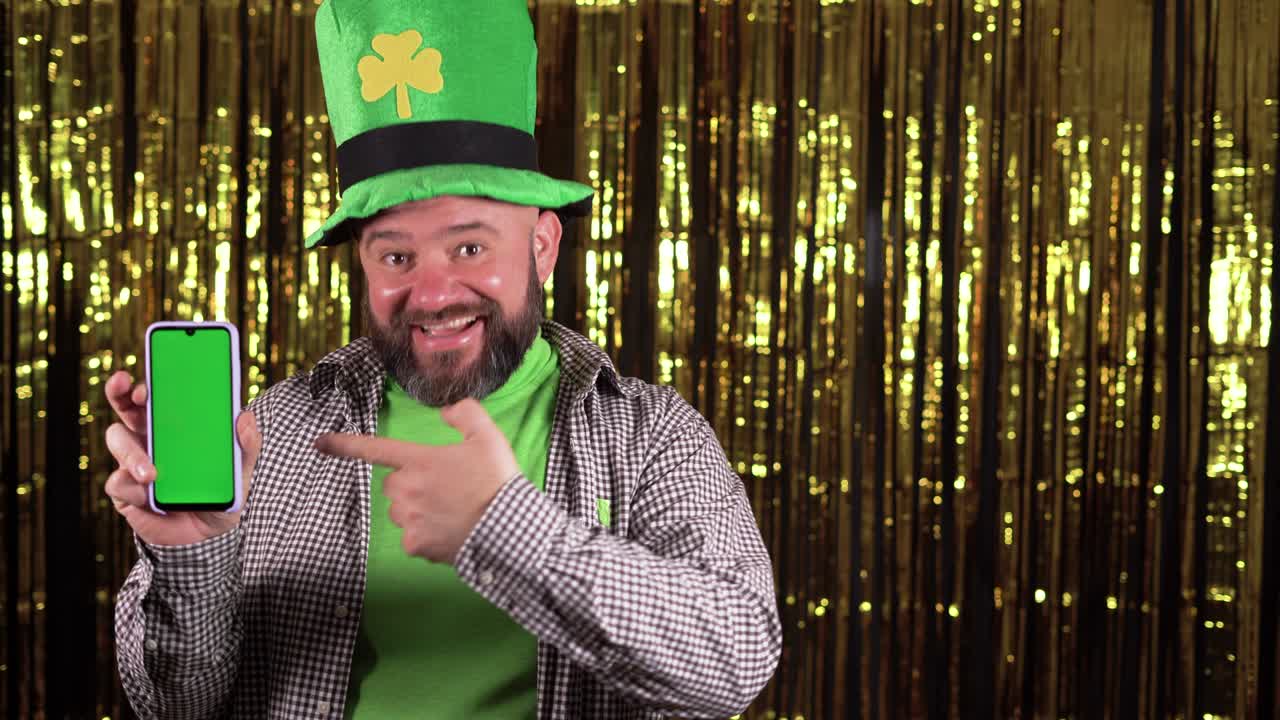 male lepricon wearing a large green shamrock hat celebrating st patrick's day. Pointing at the blank screen of your smartphone.