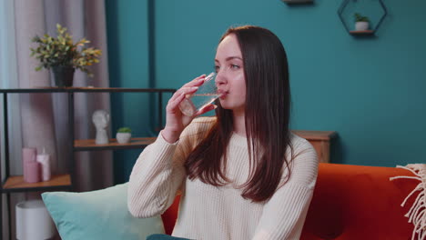 Portrait-of-young-woman-sitting-on-sofa-at-home-holds-glass-drinks-water,-makes-gulp-of-clean-aqua