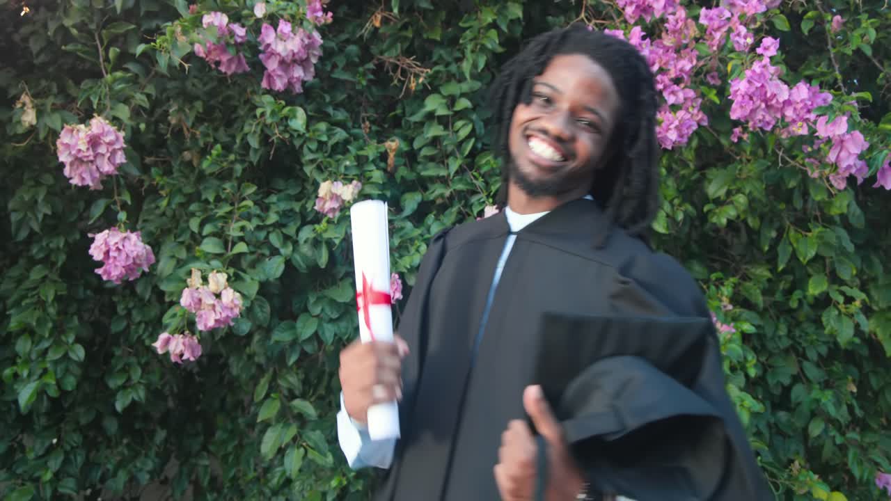 African American man celebrating in gown after getting red-ribbon diploma, waving cap in garden