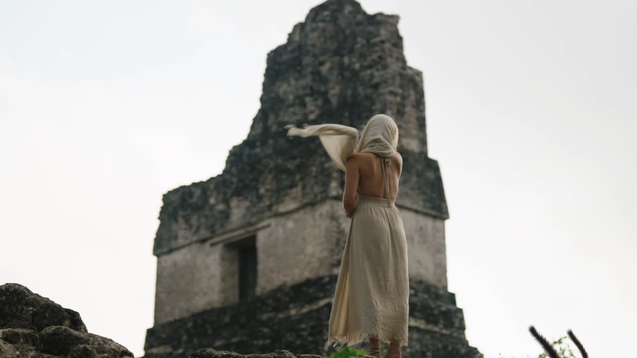 A woman in a white dress stands barefoot facing a towering Mayan pyramid at Tikal, Guatemala.