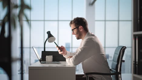 Junger-Mann,-Der-Mit-Dem-Handy-Spricht-Und-Sich-Notizen-Macht,-Indem-Er-Im-Online-Chat-Per-Handy-SMS-Schreibt.-Lächelnder-Mann-Sitzt-Am-Schreibtisch-Mit-Laptop-Im-Heimbüro-Und-Trägt-Ohrhörer