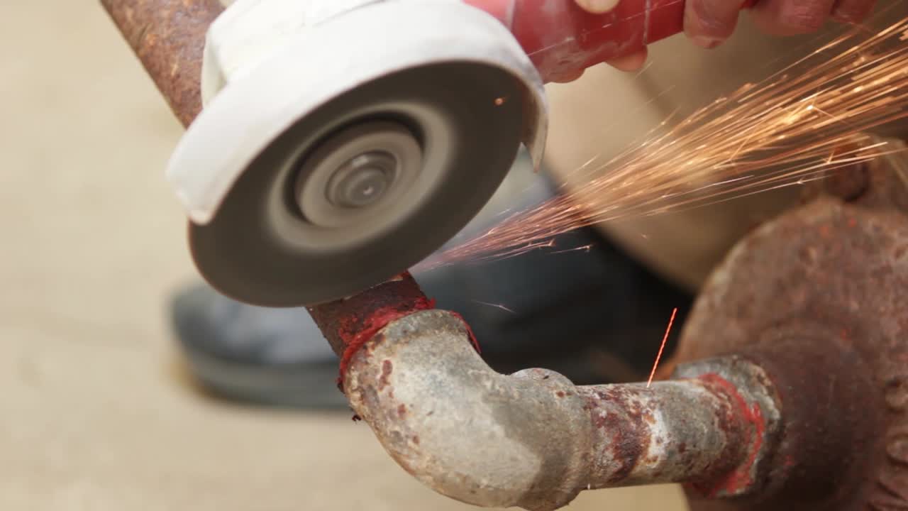 a side view of a man picking up a portable grinding machine to cut a rusty metal pipe, bright sparks