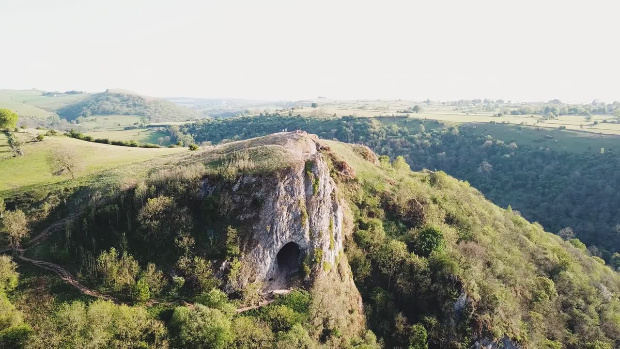 Anti-clockwise aerial drone shot of Thor's Cave, Ashbourne, Peak District at sunset. Forest, green rolling fields and clear skies in background.