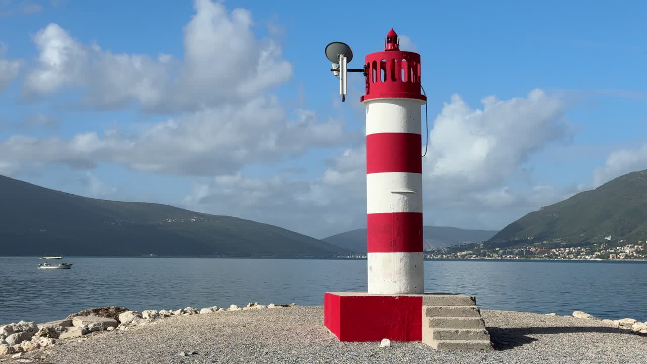 Close-up view of a red-and-white lighthouse rising above calm coastal waters under a bright blue sky with scattered clouds. Ideal for navigation, maritime travel, seascapes, coastal landscapes