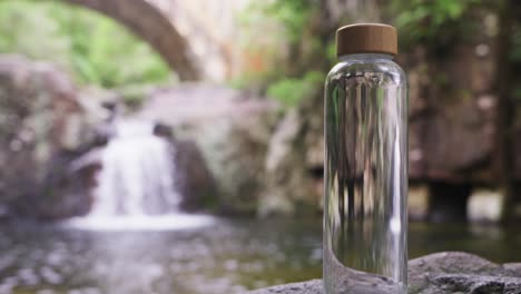 close up, pan out, a clear glass water bottle sitting on top of a rock with a flowing waterfall and rock bridge in the background.