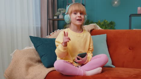Child-girl-kid-with-smartphone-in-headphones-dancing-singing-listening-music-at-home-alone-on-sofa