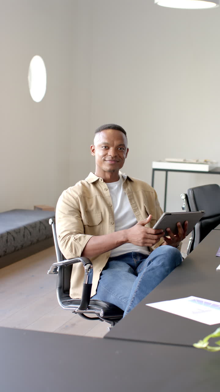 Vertical video: Sitting in office, man holding tablet and looking at camera confidently