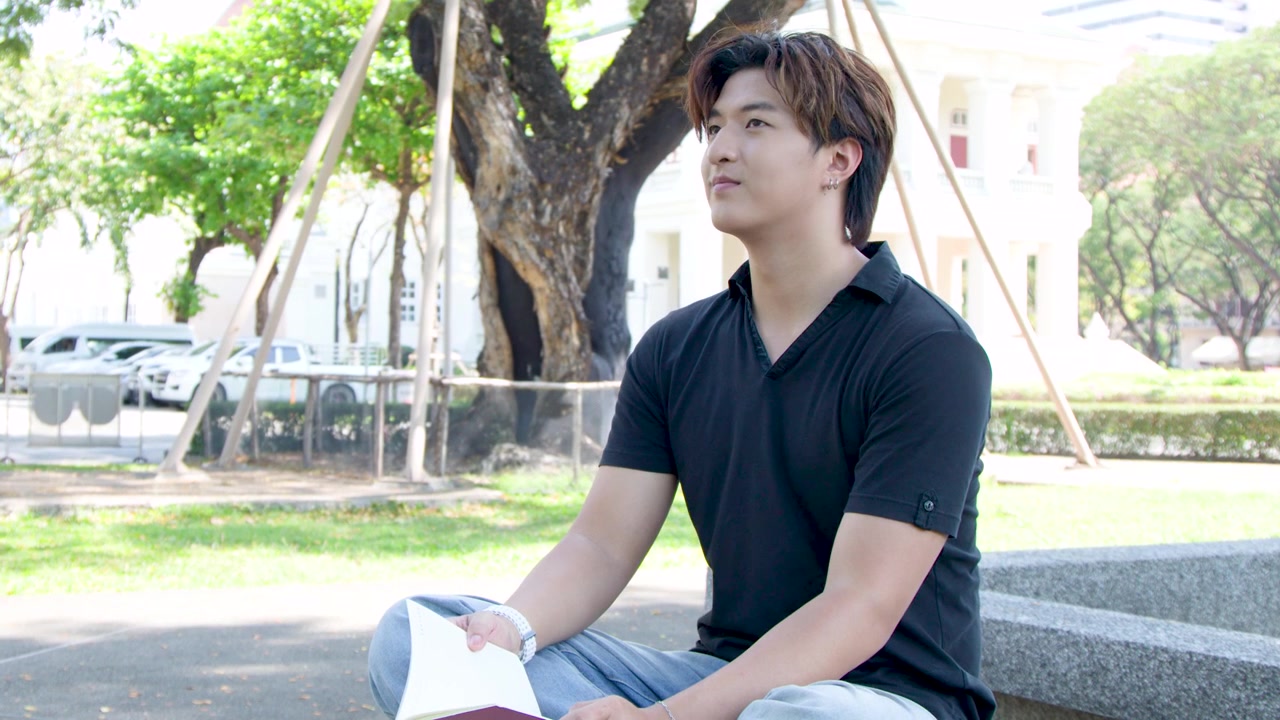 黒いシャツを着た若いタイ人男性が、屋外に座って本を読んでいる。