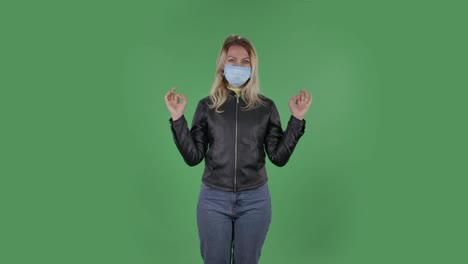 의료 보호 얼굴 마스크를 입은 아름다운 젊은 여성의 초상화는 카메라를보고 휴식을 취하고 명상하고 있습니다. 검은 재과 청바지를 입은 느슨한 머리카락을 가진 금발은 스튜디오에서 녹색 화면에 있습니다. 건강 보호 코로나 바이러스 개념