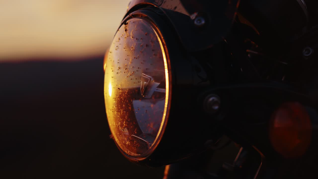 A static close-up of a motorcycle headlight glass, covered in dead insects, reflecting the warm, hazy golden hour light of a vibrant sunset. Suggests long road trip and adventure