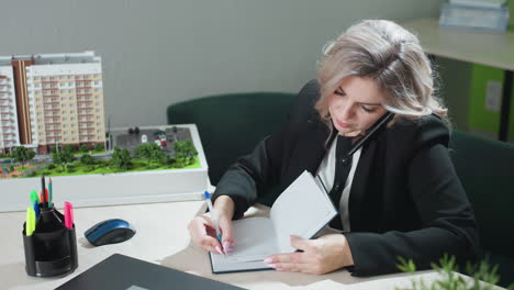 real estate agent multitasking at desk holding phone between ear and shoulder while writing in notebook with pink nails, seated in office with building model and laptop visible on workspace