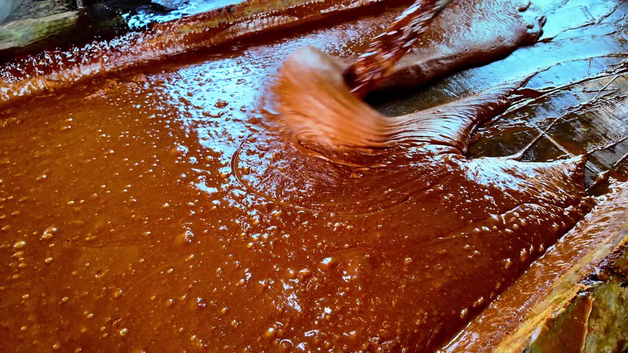 Caramelized sugar thickens in large wooden vat, stirred by hand in rustic setting